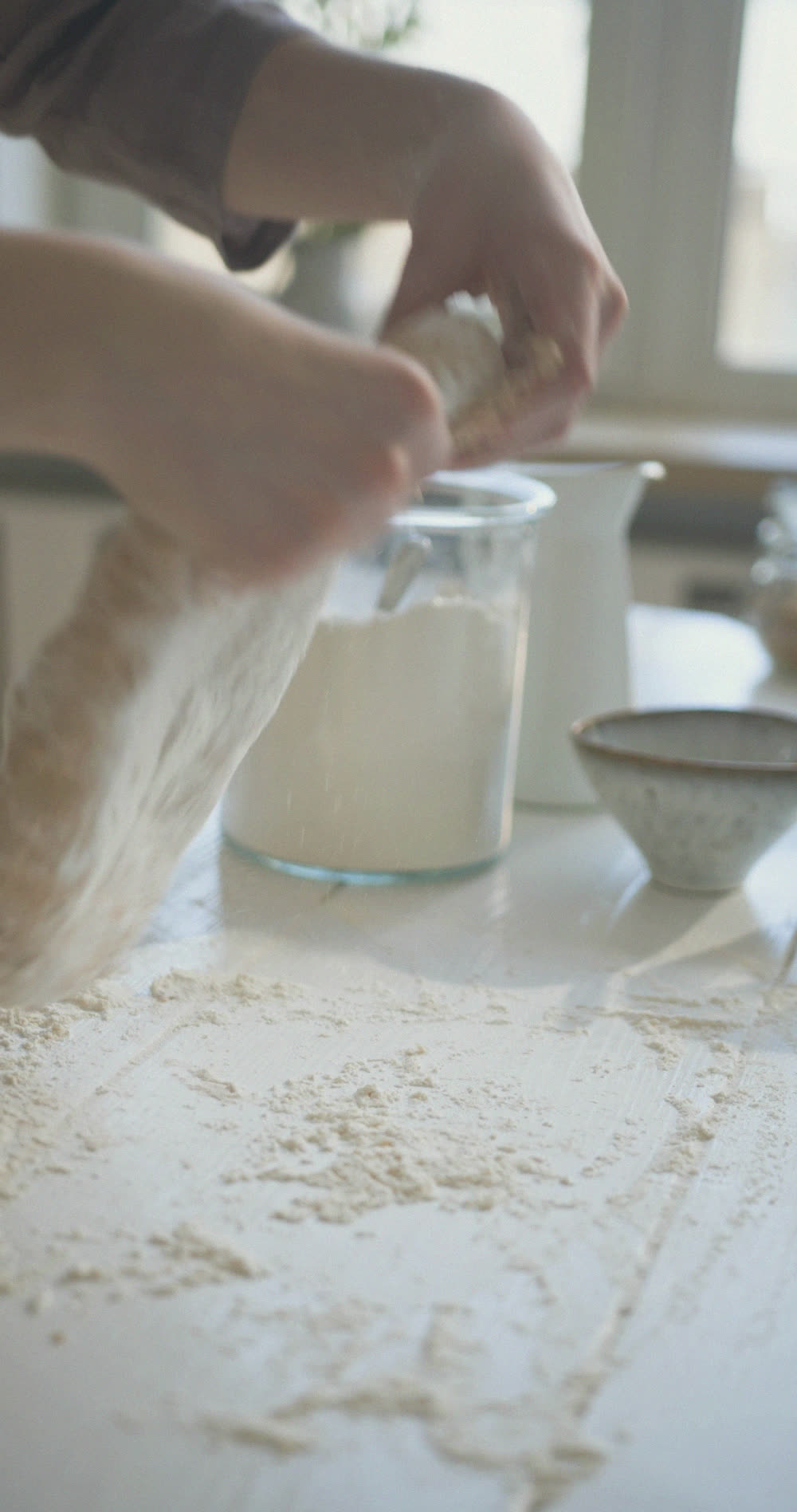 Two pairs of hands working with flour on a kitchen table, by a window.
